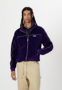 Lilla velour zip-up jakke med hvite detaljer, med høy krage og logo. Bærendes over en svart skjorte og beige bukser.
