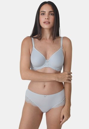 Frau mit langen dunklen Haaren, die einen hellgrauen BH und passende Unterwäsche mit Spitzenbesatz trägt, steht vor einem schlichten Hintergrund.