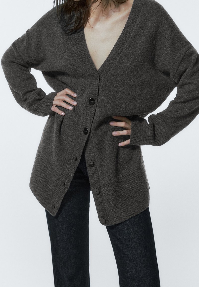 Femme portant un cardigan gris foncé surdimensionné boutonné et un pantalon noir, mains sur les hanches, visible du cou aux cuisses.