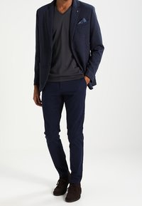 Granatowa marynarka o fakturze, nałożona na ciemnoszarą koszulkę z dekoltem w serek, zestawiona z granatowymi chinosami i ciemnobrązowymi butami, z poszetką w kieszeni.