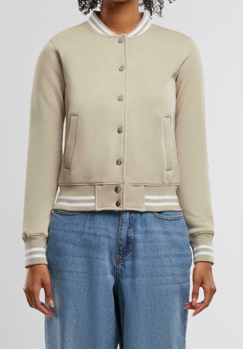 Personne portant une veste bomber beige boutonnée avec des poignets et un col rayés blancs, associée à un jean bleu, debout devant un fond uni.