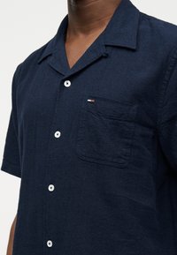 Σκούρο μπλε πουκάμισο με κοντά μανίκια και κουμπιά, με μικρή τσέπη στο στήθος και λευκά κουμπιά, φορεμένο από άτομο που απεικονίζεται από το λαιμό μέχρι τη μέση.