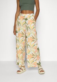 Femme portant un pantalon à jambes larges avec un motif floral orange et vert, un crop top vert olive et des sandales beiges, debout devant un fond blanc.