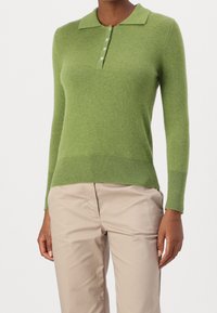 Kobieta ubrana w długi rękaw, jasnozielony sweter polo z dzianiny, z czterema guzikami i beżowymi spodniami, pokazana od szyi do połowy uda.