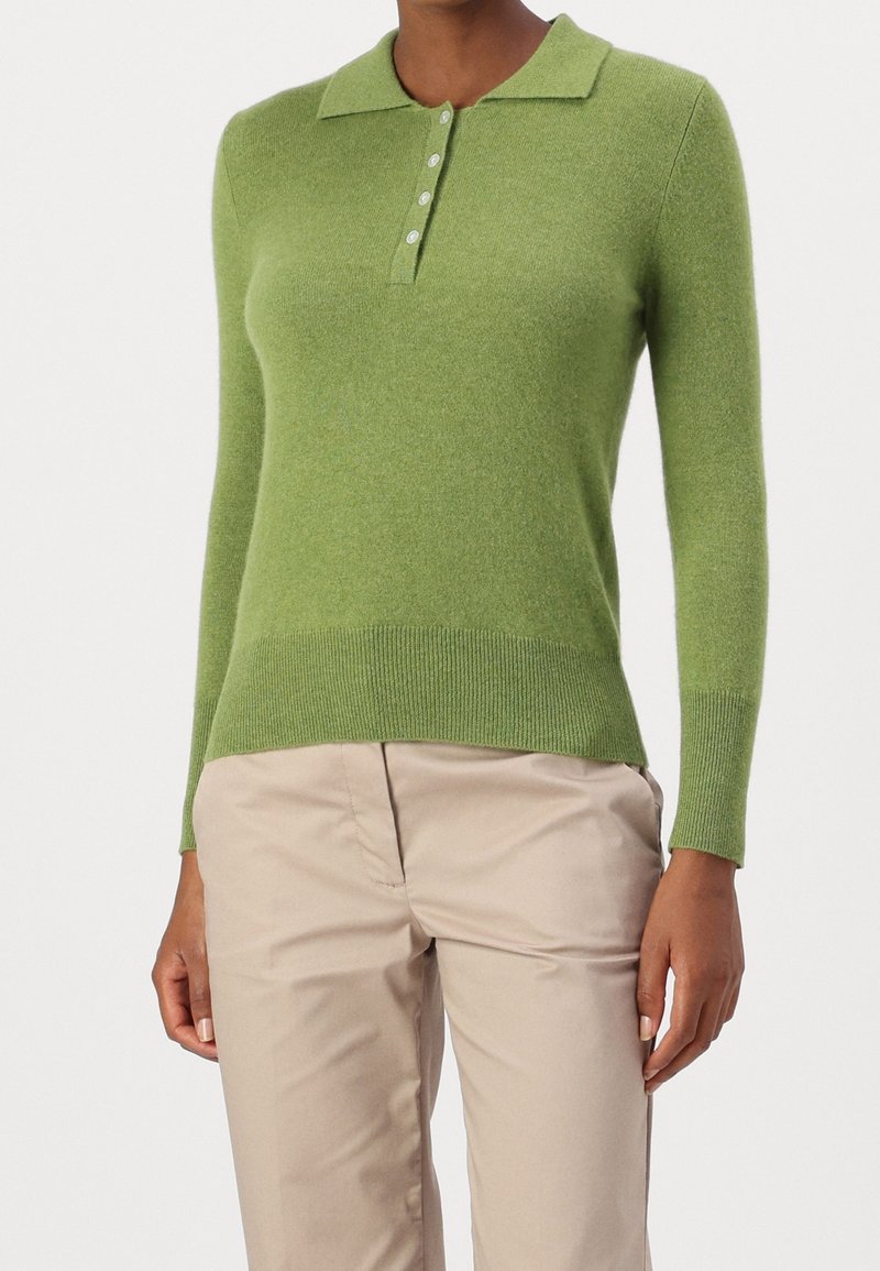 Kobieta ubrana w długi rękaw, jasnozielony sweter polo z dzianiny, z czterema guzikami i beżowymi spodniami, pokazana od szyi do połowy uda.