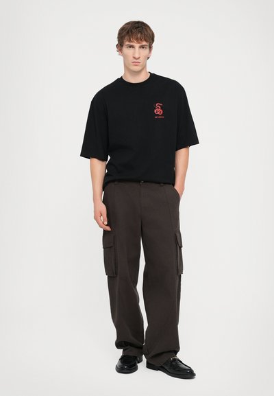 Schwarzes Baumwoll-T-Shirt mit rotem Grafiklogo, kombiniert mit dunkelgrauen Cargo-Hosen mit Seitentaschen und lässigem Schnitt, ergänzt durch schwarze Schuhe.