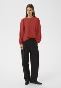 Femme debout portant un chemisier rouge à manches longues ample, un pantalon noir à jambes larges et des chaussures noires sur un fond gris clair uni.