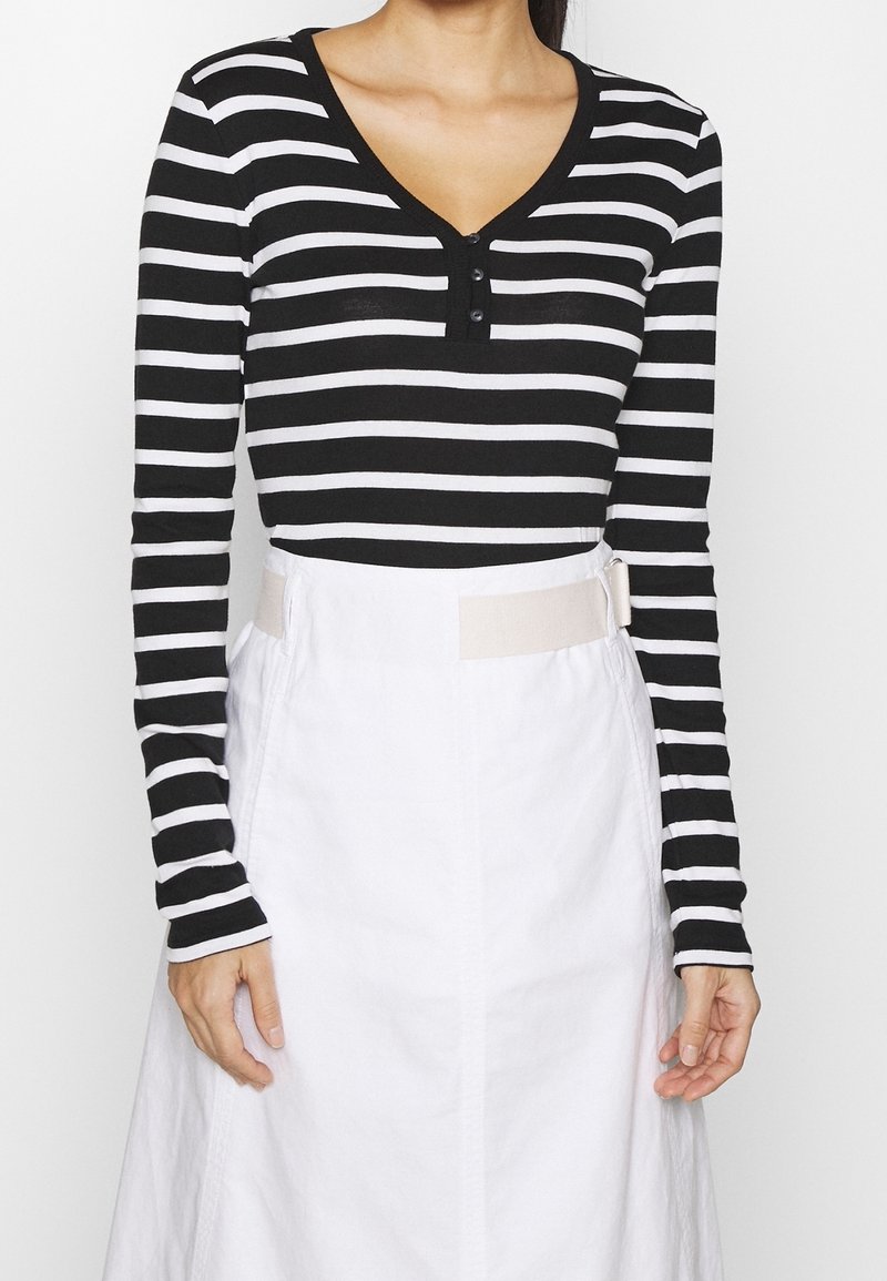 Woman wearing a black and white horizontal striped long-sleeve top with buttons, paired with a white high-waisted skirt with side straps.