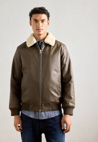 Giacca in pelle marrone con colletto in ecopelliccia color crema, zip frontale, polsini a coste e due tasche laterali, indossata sopra una camicia a scacchi.
