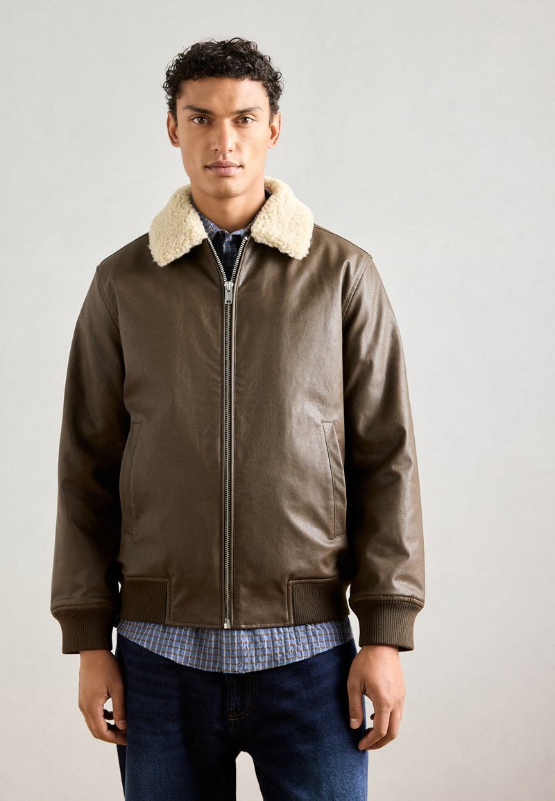 Giacca in pelle marrone con colletto in ecopelliccia color crema, zip frontale, polsini a coste e due tasche laterali, indossata sopra una camicia a scacchi.