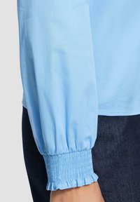 Blusa azzurro chiaro con polsini arricciati ed elasticizzati. Tessuto liscio e intrecciato, maniche lunghe e una vestibilità comoda. Abbinata a jeans scuri.