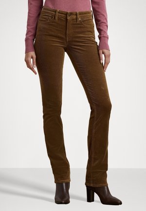 Vrouw met bruine corduroy broek en donkerbruine enkellaarsjes met hak, met een mauve lange mouwen top, staand tegen een effen achtergrond.
