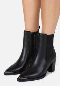 Botas de cuero negro hasta el tobillo con punteras puntiagudas, paneles laterales elásticos, cierres de cremallera y acentos metálicos en la punta. Tacones altos y gruesos.