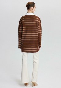 Bruin gestreepte lange mouw trui met een ontspannen fit, gecombineerd met witte flared broek en beige hakken, gezien van achteren.