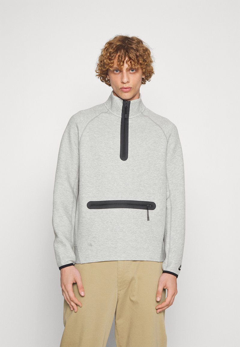 Pull en polaire gris clair avec un col montant, fermeture éclair noire et poche zippée à l'avant. Il dispose de manches raglan et d'accents noirs contrastants.