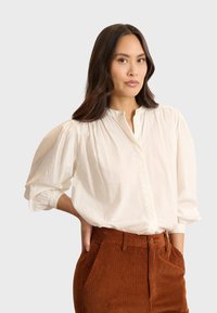 Camicia bianca con bottoni a maniche a sbuffo, dotata di patta e texture morbida, abbinata a pantaloni in velluto a coste color ruggine.