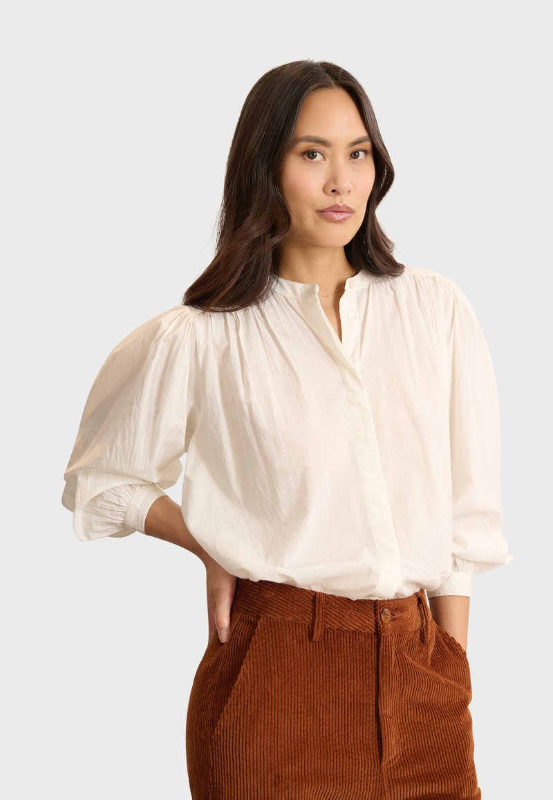 Camicia bianca con bottoni a maniche a sbuffo, dotata di patta e texture morbida, abbinata a pantaloni in velluto a coste color ruggine.