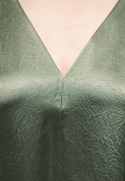 Κοντινό πλάνο σε τσαλακωμένο, ελιά πράσινο ύφασμα με V-λαιμόκοψη, με ορατή ραφή και ομαλή ανοιχτόχρωμη περιοχή δέρματος στο πάνω κέντρο.