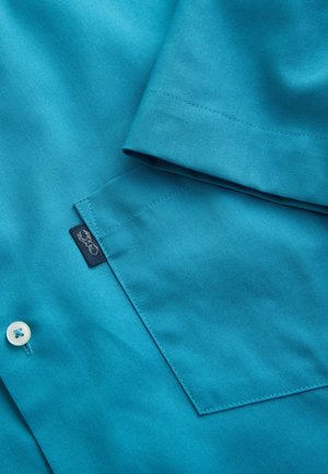 Nahaufnahme einer türkisblauen Hemdtasche mit ordentlichen Nähten, einem weißen Knopf darunter und einem kleinen schwarzen Stoffetikett mit einem weißen Hirschlogo.