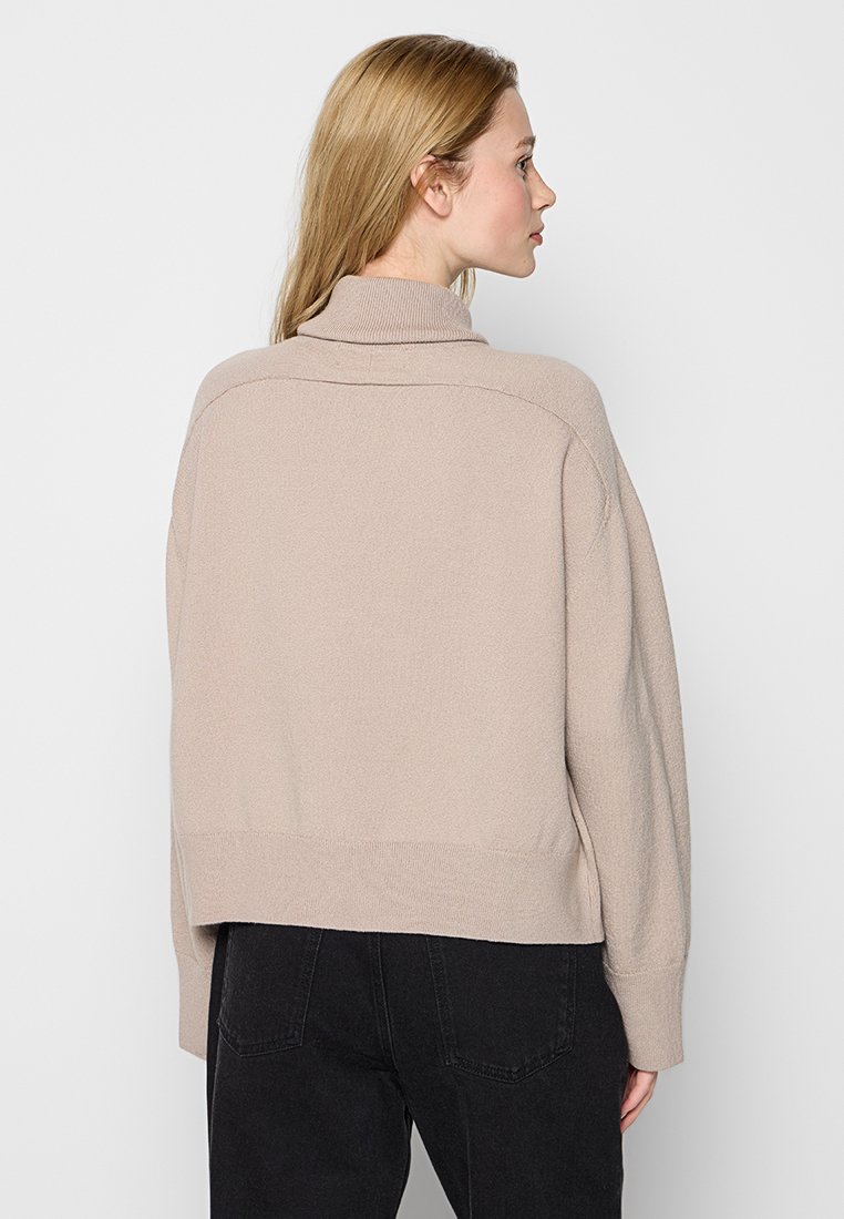 Frau mit blonden Haaren, die einen beigen Rollkragenpullover und schwarze Jeans trägt und seitlich steht, vor einem neutralen Hintergrund.
