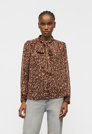 Femme aux cheveux tressés portant un chemisier marron à imprimé léopard avec un nœud au col et un jean gris clair, debout devant un fond uni.