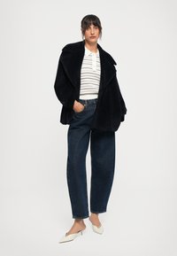 Casaquinho preto e peludo de tamanho exagerado, camisa polo branca com riscas, calças de ganga escuras com corte relaxado, mules bejes com biqueira fina e salto baixo.