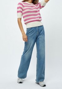 Žena oblečená v pleteném topu s růžovými a bílými pruhy, v modrých džínech s vysokým pasem a širokými nohavicemi, a v bílých teniskách, stojí s jednou rukou vztyčenou.