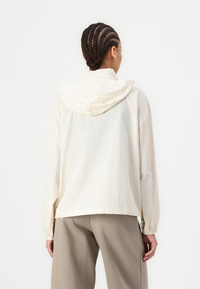 Veste à capuche crème en matériau léger, offrant une coupe décontractée, des poignets élastiques et une texture lisse. Finitions soignées des coutures à l'ourlet.