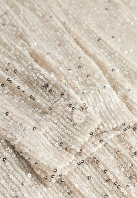 Bežinė blizgučių medžiaga su mažais juodais akcentais, turinti mygtukų užsegimą ir lygų paviršių. Blizgučiai sukuria mirgantį efektą.