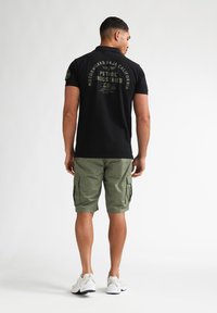Czarna koszulka polo z zielonymi grafikami na plecach, w połączeniu z oliwkowozielonymi szortami cargo i białymi butami sportowymi. Gładka tkanina, luźny fason.