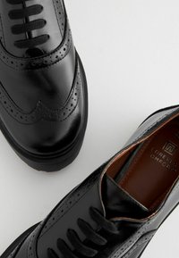Zapatos de cuero negro pulido con detalles brogue, punta redonda, cierre de cordones y suela de goma texturizada. El interior presenta un forro marrón.