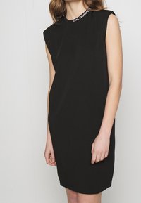 Czarna sukienka bez rękawów z okrągłym dekoltem, z białym akcentem marki przy kołnierzyku. Gładki materiał, luźny krój i długość do kolan.