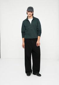 Felpa con zip di un verde scuro con logo "Originals", abbinate a pantaloni larghi neri e un cappellino grigio, in piedi su uno sfondo chiaro.