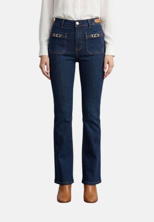 Jeans blu scuro a gamba dritta con tasche applicate sul davanti con piccoli dettagli in catena metallica, indossati con stivaletti marroni e una camicetta bianca.