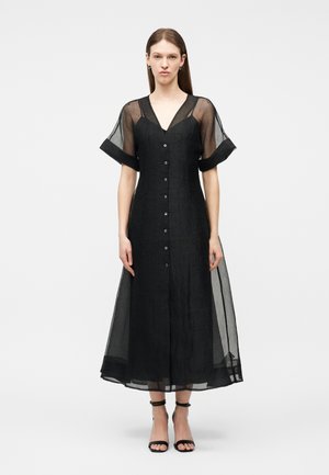 Femme portant une robe midi noire boutonnée avec des manches courtes transparentes et un ourlet transparent, associée à des talons noirs ouverts à la cheville, debout devant un fond blanc.