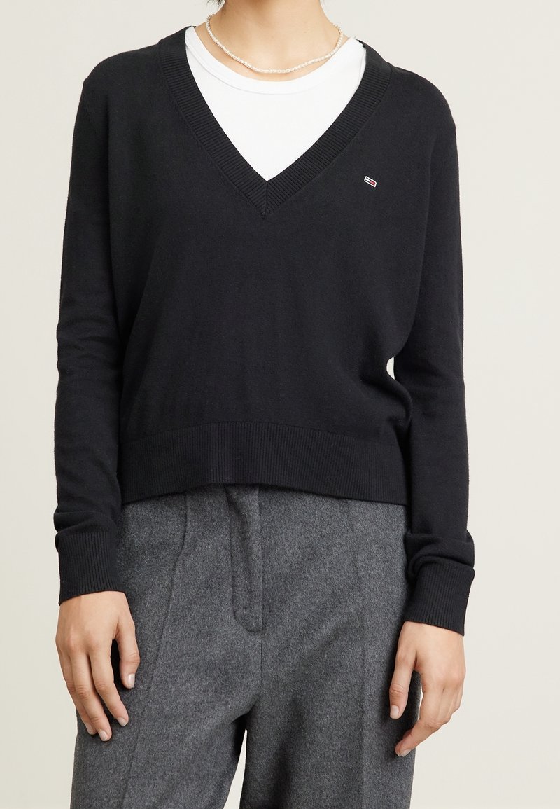 Personne portant un pull noir à col en V sur une chemise blanche et un pantalon gris, accessoirisée d'un court collier de perles.