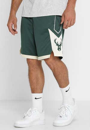 Männliche Beine, die grüne Basketballshorts mit einem weißen Hirschlogo, weiße Nike-Socken und weiße Nike-Sneakers tragen.