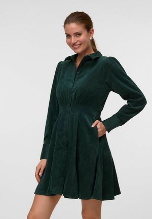 Femme souriante portant une robe boutonnée à manches longues vert foncé avec une taille cintrée et une jupe évasée, une main dans la poche.