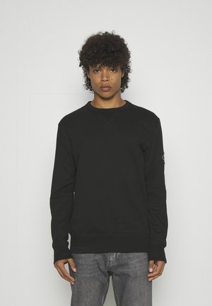 Mladá osoba s kudrnatými vlasy nosící obyčejnou černou mikinu s dlouhým rukávem a šedé džíny na jednoduchém bílém pozadí.