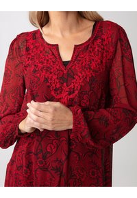 Blouse rouge brodée de fleurs avec des motifs noirs, présentant un décolleté en V et des manches en cloche. Le tissu texturé ajoute de la profondeur au design.