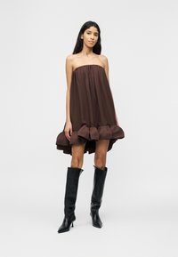 Γυναίκα που φορά ένα στράπλες, φαρδύ καφέ φόρεμα με βολάν στο τελείωμα και μαύρες ψηλές μπότες με τακούνι έως το γόνατο, στέκεται μπροστά σε απλό φόντο.