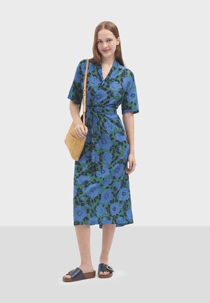 Robe portefeuille à fleurs avec des manches courtes ; motif bleu et vert ; taille à nouer ; associée à des sandales noires à glissière ; le mannequin tient un sac en paille texturé.