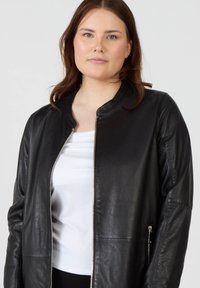 Svart läderjacka med en slät textur, rund halsringning, framzipper och sidfickor. Bärs över en vit topp.