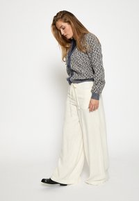 Siva in bež pletena bunda z teksturiranim vzorcem, v kombinaciji s širokimi belimi hlačami in črnimi čevlji. Ženska stoji v bočnem profilu.