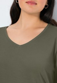 Olivgrünes, kurzärmeliges V-Ausschnitt-Top aus weichem Stoff, mit glatter Textur und sauberem Stitching entlang des Ausschnitts.
