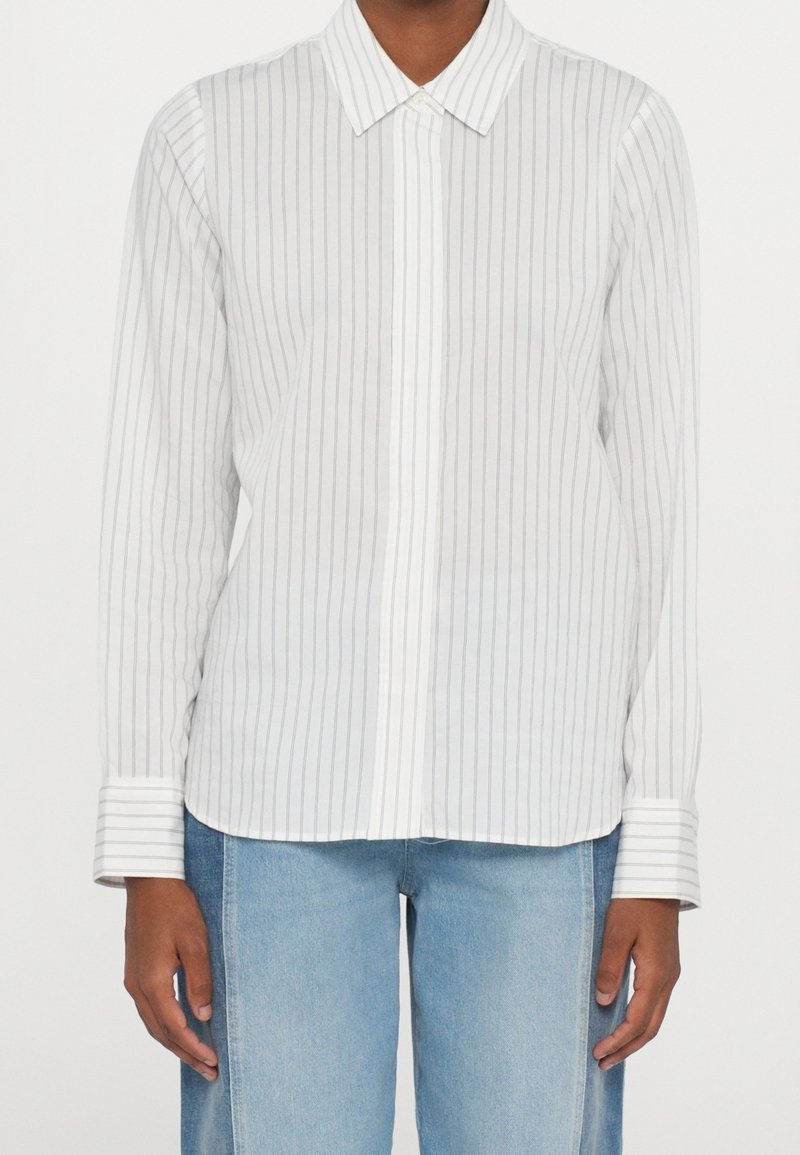 Personne portant une chemise blanche à manches longues avec de fines rayures verticales grises et un jean bleu clair, debout les bras relâchés le long du corps.