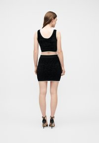  Γυναίκα όρθια, φοράει μαύρο αμάνικο κοντό τοπ και φούστα ψηλόμεση σετ, με μαύρες ψηλοτάκουνες γόβες, γυρισμένη πλάτη.