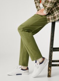 Pantaloni din bumbac verde cu o formă dreaptă, asortați cu adidași albi cu accente maro și bleumarin. Picioarele încrucișate, așezat pe un taburet negru.
