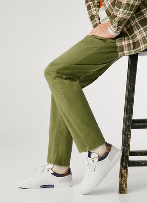 Green cotton trousers with a straight shape, paired with white sneakers featuring brown and navy accents. Legs crossed, seated on a black stool.