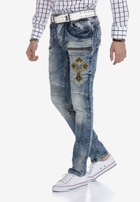 Ausgebleichte blaue Jeans mit dekorativen Nähten, einem Kreuzsymbol mit Verzierungen und einem slim fit Design. Kombiniert mit weißen Sneakers.